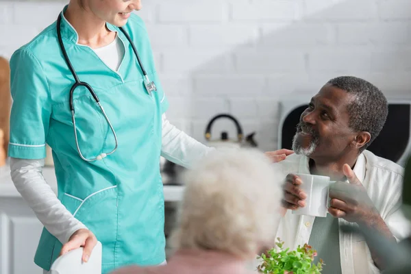 Africano americano uomo tenendo tazza vicino sorridente infermiera in casa di cura — Foto stock