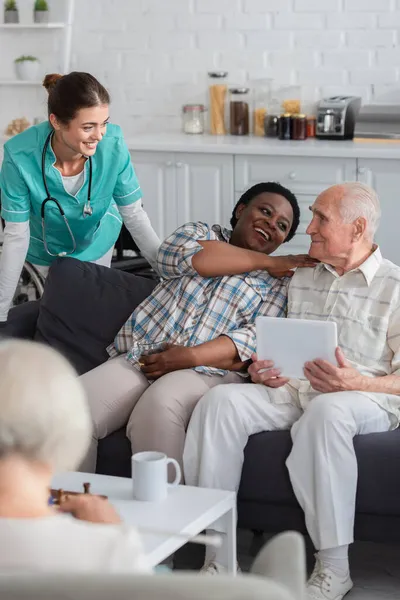 Glückliche Afroamerikanerin mit Tee schaut Freundin mit digitalem Tablet nahe Krankenschwester im Pflegeheim an — Stockfoto