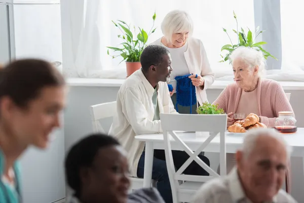 Улыбающиеся межрасовые пациенты с пряжей и чаем разговаривают в доме престарелых — стоковое фото