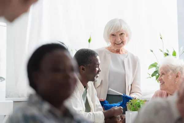 Elderly woman holding yarn near multiethnic pensioners in nursing home — Stock Photo