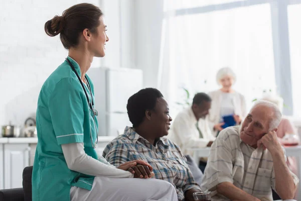 Enfermera joven cogida de la mano de un paciente afroamericano con té en un asilo de ancianos - foto de stock