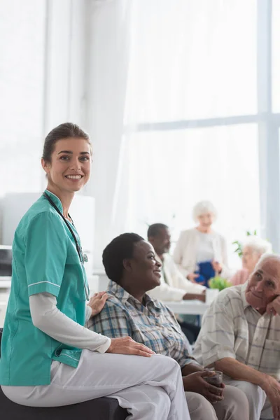 Jeune infirmière souriant à la caméra près de patients multiethniques dans une maison de retraite — Photo de stock