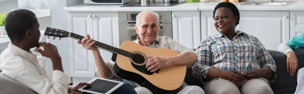 Hombre mayor tocando la guitarra acústica cerca de amigos multiétnicos con tableta digital en el hogar de ancianos, pancarta - foto de stock
