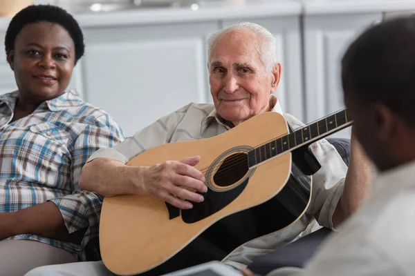 Hombre mayor tocando la guitarra acústica cerca de amigos afroamericanos en un asilo de ancianos - foto de stock