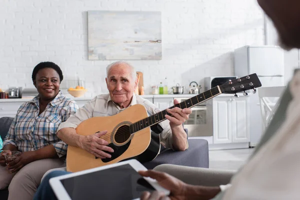Багатоетнічні літні люди з акустичною гітарою та цифровим планшетом у будинку престарілих — стокове фото