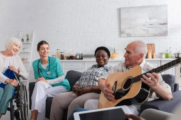 Старший мужчина держит акустическую гитару рядом с многонациональными друзьями и медсестрой в доме престарелых — стоковое фото