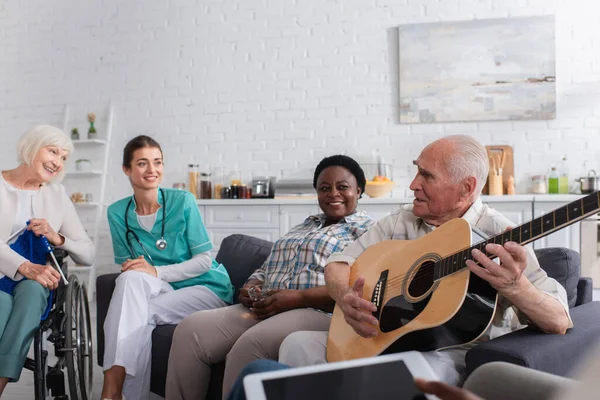 Homem sênior segurando guitarra acústica perto de amigos multiétnicos e enfermeira em casa de repouso — Fotografia de Stock