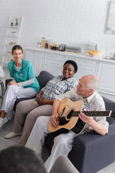 Homem idoso tocando guitarra acústica perto de amigo afro-americano com chá e enfermeira em casa de repouso — Fotografia de Stock