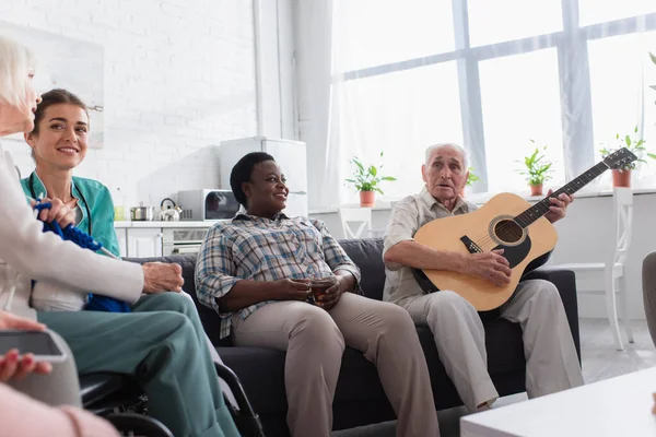 Многонациональные пожилые люди с акустической гитарой и пряжей проводят время рядом с медсестрой в доме престарелых — стоковое фото