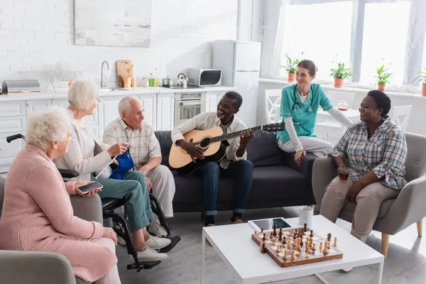 Hombre afroamericano tocando la guitarra acústica cerca de amigos con smartphone, hilo y enfermera en el hogar de ancianos - foto de stock