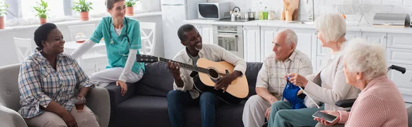 Smiling interracial elderly people playing acoustic guitar near nurse in nursing home, banner — Stock Photo