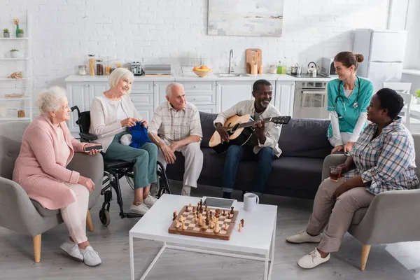 Multiethnic senior people with acoustic guitar, smartphone and yarn spending time near nurse in nursing home — Stock Photo