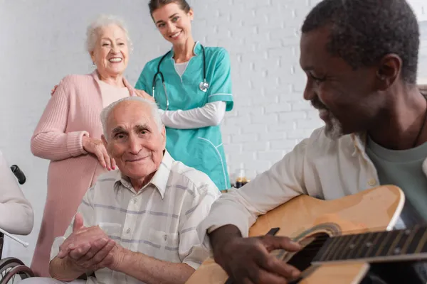 Старший мужчина сидит рядом с межрасовыми друзьями с акустической гитарой и медсестрой в доме престарелых — стоковое фото