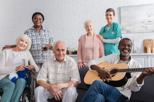 Pensionistas interracial positivos con hilo y guitarra acústica mirando a la cámara cerca de la enfermera en el hogar de ancianos - foto de stock