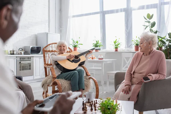 Mujer mayor feliz tocando la guitarra acústica cerca de amigos multiétnicos con dispositivos y ajedrez en el hogar de ancianos - foto de stock