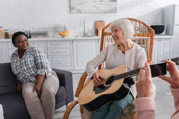 Пожилая женщина играет на акустической гитаре рядом с межрасовыми друзьями с мобильным телефоном в доме престарелых — стоковое фото