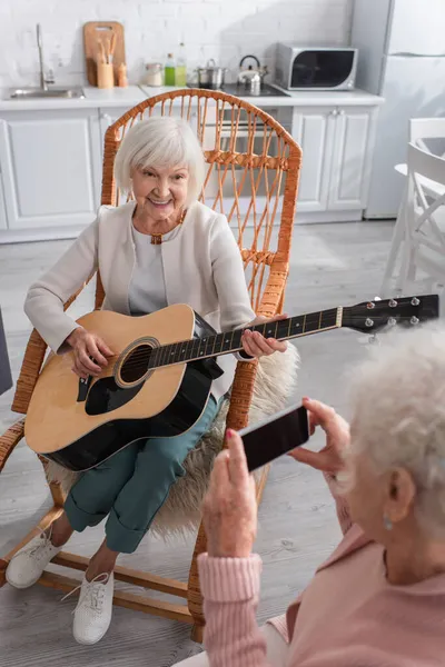 Femme âgée souriante jouant de la guitare acoustique près d'un ami avec smartphone dans une maison de retraite — Photo de stock