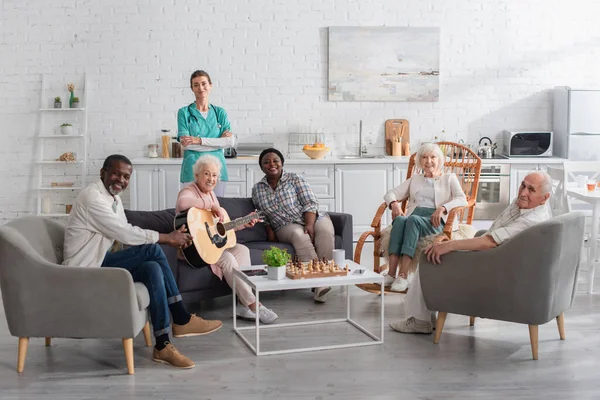 Cheerful interracial people with acoustic guitar looking at camera near chess and nurse in nursing home — Stock Photo