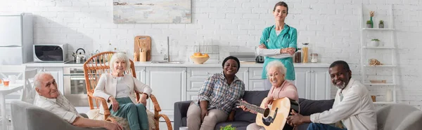 Positive multiethnische Senioren mit Akustikgitarre, die in der Nähe einer Krankenschwester im Pflegeheim in die Kamera schauen, Banner — Stockfoto