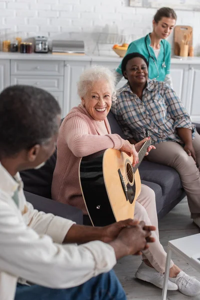 Mujer anciana sonriente tocando la guitarra acústica cerca de amigos multiétnicos y enfermera borrosa en el hogar de ancianos - foto de stock