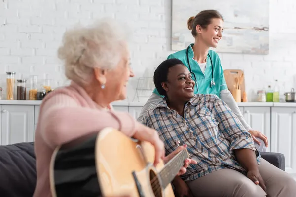 Enfermera sonriente y mujer afroamericana sentada cerca de paciente borrosa tocando guitarra acústica en hogar de ancianos - foto de stock
