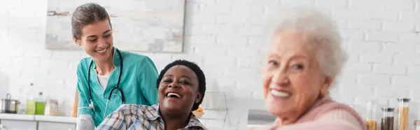 Sonrientes mujeres interracial mirando a la cámara cerca de la enfermera en el hogar de ancianos, pancarta - foto de stock