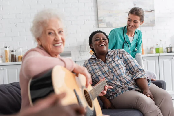 Позитивные межрасовые женщины с акустической гитарой смотрят в камеру рядом с медсестрой в доме престарелых — стоковое фото