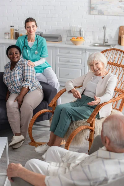 Багатоетнічні літні люди розмовляють біля медсестри на дивані в будинку престарілих — стокове фото