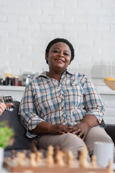 Piacevole donna afroamericana guardando la fotocamera vicino amico con chitarra acustica e scacchi in casa di cura — Foto stock