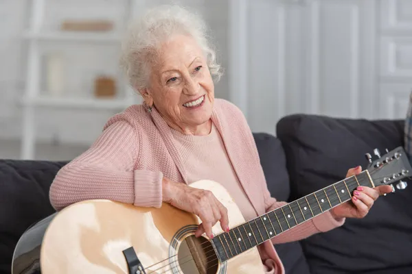 Веселая пожилая женщина играет на акустической гитаре на диване в доме престарелых — стоковое фото