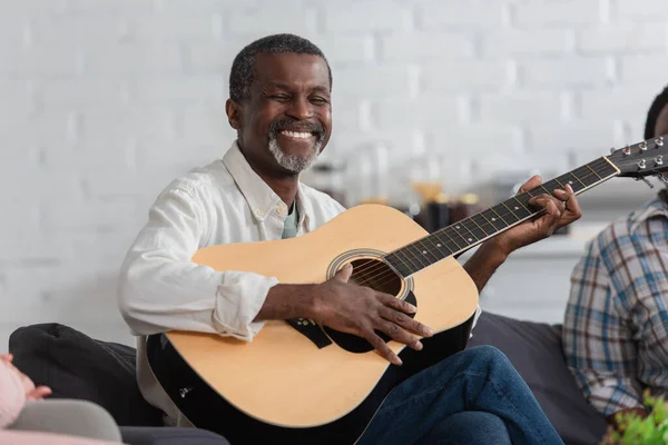 Feliz homem americano africano sênior tocando guitarra acústica no sofá em casa de repouso — Fotografia de Stock