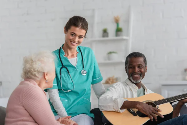 Enfermera positiva pasando tiempo con pacientes mayores interracial con guitarra acústica en residencia de ancianos - foto de stock