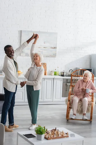 Pensionistas multiétnicos sonrientes bailando cerca de un amigo con té en un asilo - foto de stock