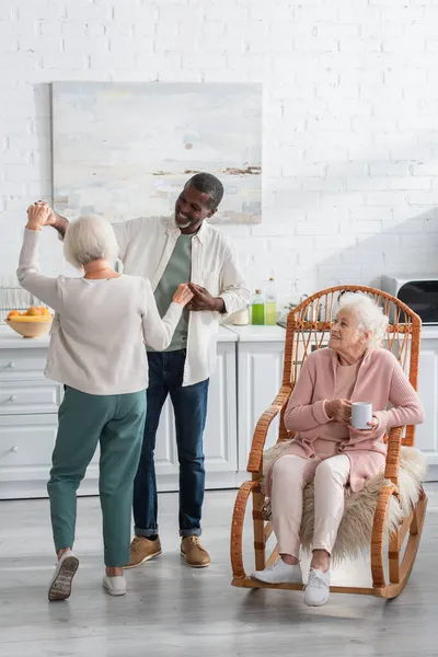 Des retraités multiethniques positifs dansent près d'un ami aîné dans une chaise berçante dans une maison de retraite — Photo de stock