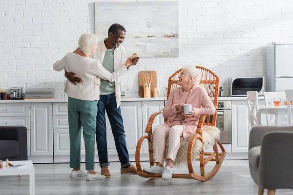 Femme âgée souriante tenant une tasse dans un fauteuil à bascule près d'amis interraciaux dansant dans une maison de retraite — Photo de stock