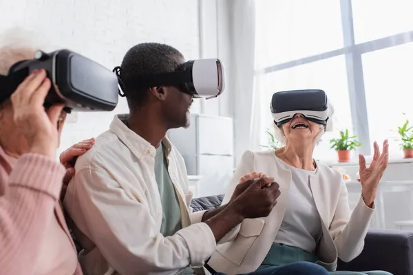 Cheerful senior woman holding hand of african american friend while gaming in vr headsets in nursing home — Stock Photo