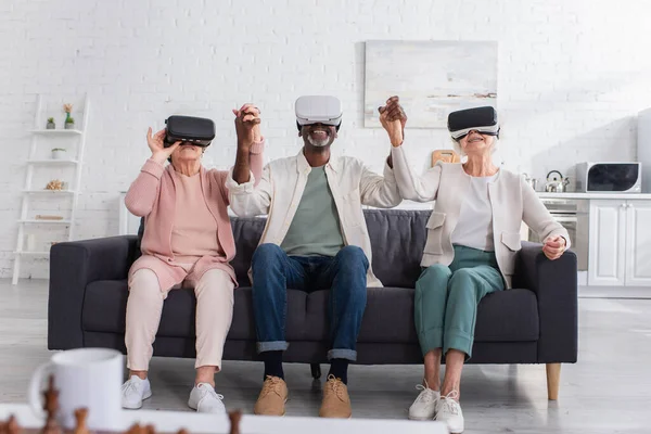 Positive multiethnic pensioners holding hands while playing in vr headsets in nursing home — Stock Photo