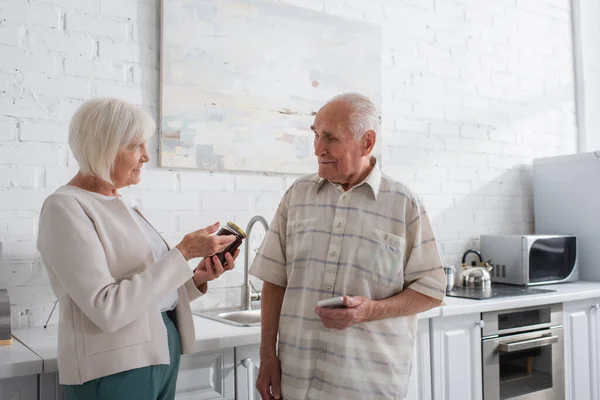 Mujer mayor sonriente señalando frasco con mermelada cerca de amigo con teléfono inteligente en el hogar de ancianos - foto de stock