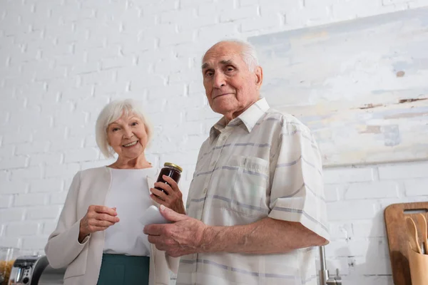 Vista ad angolo basso dell'uomo anziano che tiene lo smartphone vicino all'amico con vaso di marmellata nella casa di cura — Foto stock