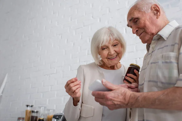 Homem idoso mostrando smartphone para amigo sorridente com frasco de geléia em lar de idosos — Fotografia de Stock