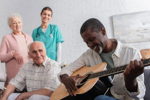 Африканский пенсионер играет на акустической гитаре рядом с друзьями и медсестрой в доме престарелых — стоковое фото