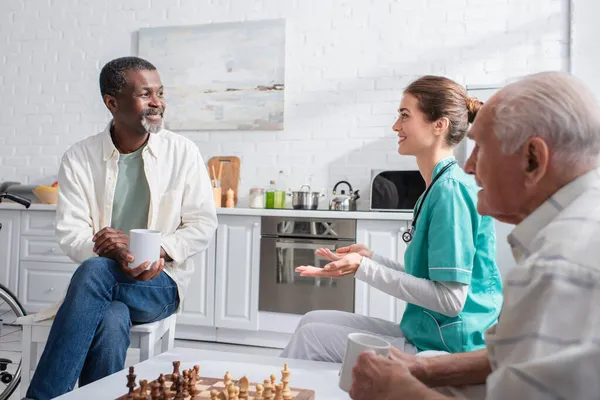 Infirmière parlant à des patients interracial avec des tasses près de l'échiquier dans une maison de retraite — Photo de stock