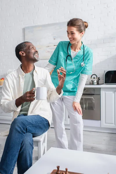 Sonriente hombre afroamericano sosteniendo taza cerca de la enfermera joven y tablero de ajedrez en el hogar de ancianos - foto de stock