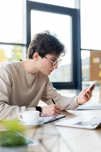 Homme d'affaires utilisant smartphone et écriture sur ordinateur portable près de tasse de café dans le bureau — Photo de stock