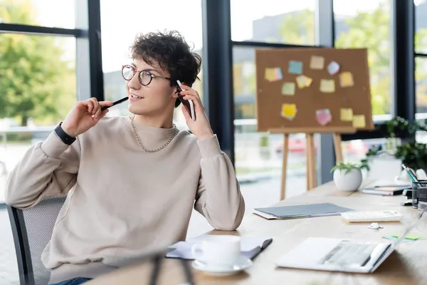 Lächelnder Geschäftsmann mit Stift und Smartphone neben Laptop, Kaffee- und Papiermappe im Büro — Stockfoto