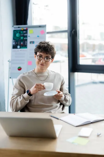 Geschäftsmann mit Brille hält Tasse und Untertasse neben verschwommenem Laptop und Notizbuch im Büro — Stockfoto