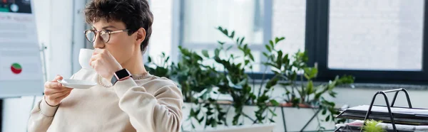 Young businessman in eyeglasses drinking coffee in office, banner — Stock Photo