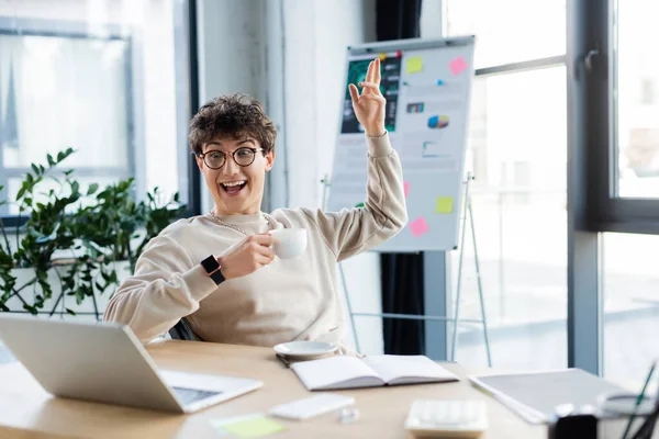 Uomo d'affari allegro con coppa che ha videochiamata su computer portatile vicino a notebook in ufficio — Foto stock