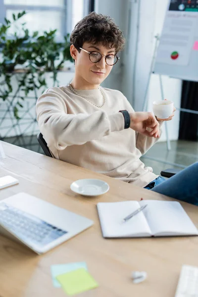 Young businessman holding cup and looking at smartwatch in office — Stock Photo