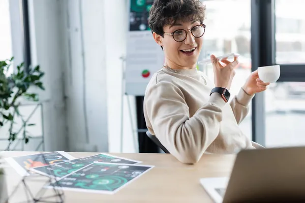 Positivo messaggio vocale uomo d'affari di registrazione su smartphone e tenendo tazza vicino laptop e carte in ufficio — Foto stock